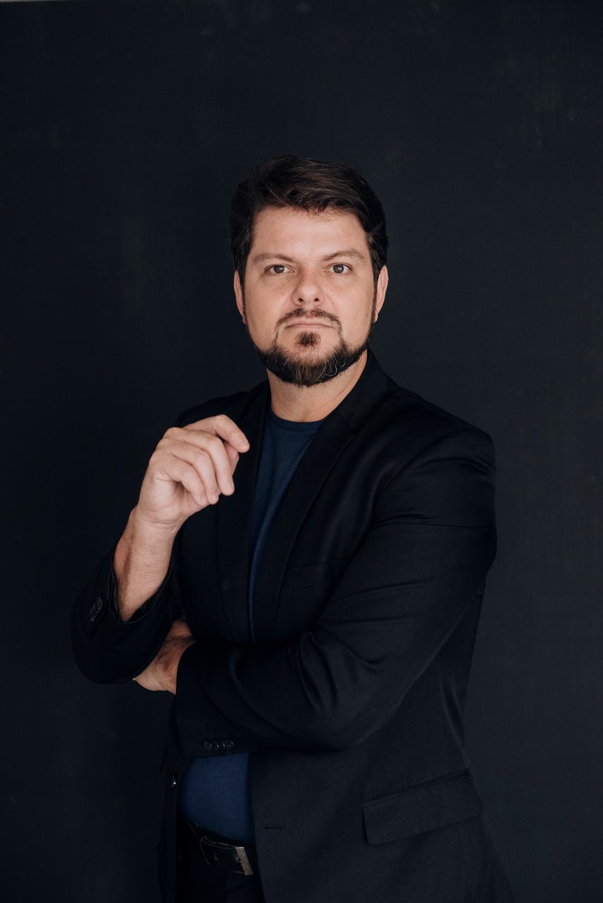 Professor Gustavo Tato, de blazer preto, em retrato sobre fundo escuro