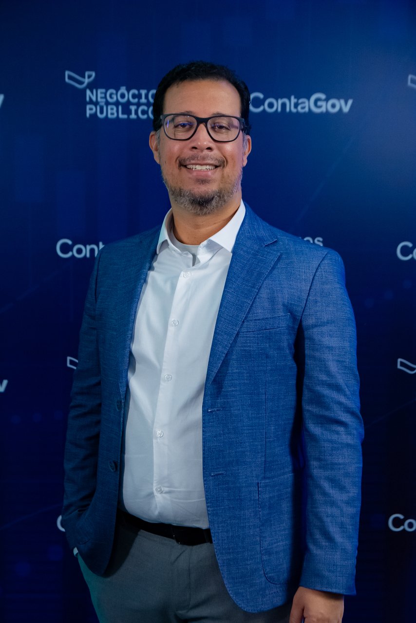 Professor Rafael Mota, de óculos, blazer azul e camisa branca, sorrindo, em retrato sobre fundo azul
