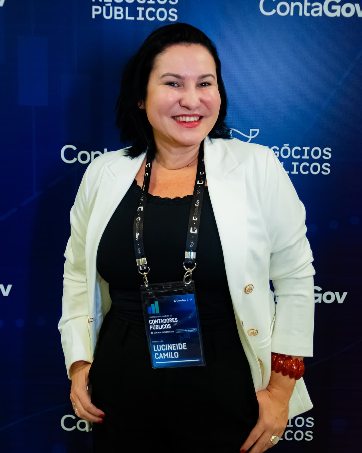 A instrutora Lucineide Camilo, sorridente, de blazer branco, em um evento da ContaGov e do Grupo Negócios Públicos